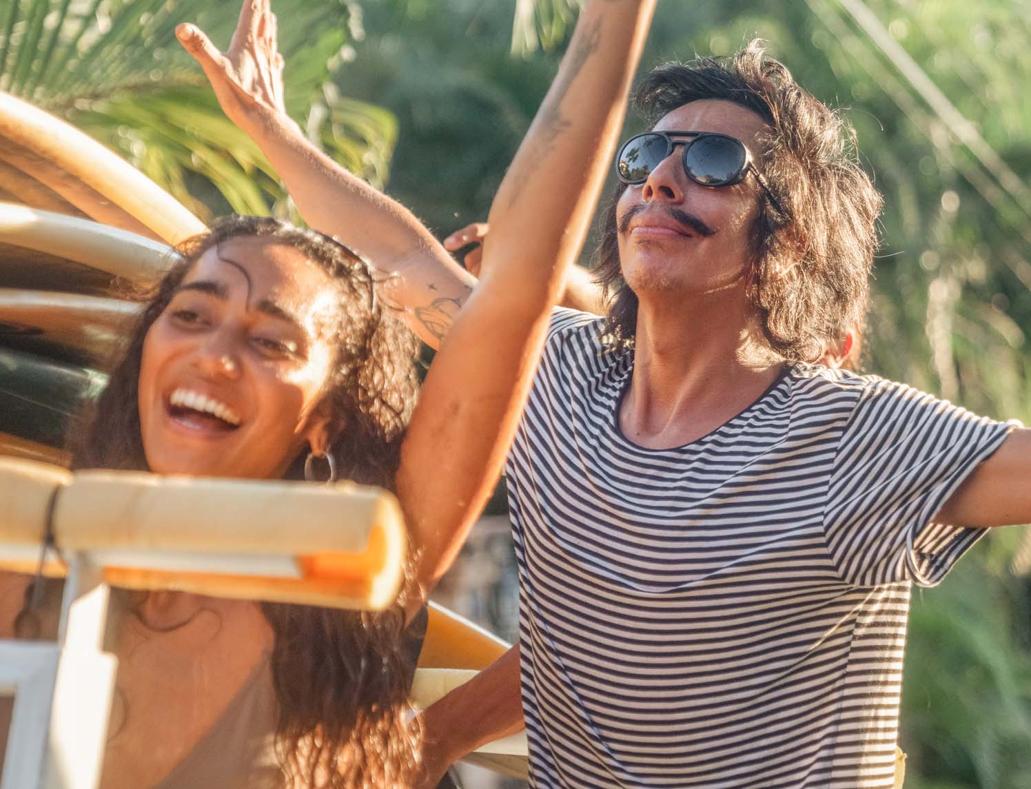 Man and woman smiling in the sun next to a car carrying surfboards wearing Ombraz armless strap sunglasses