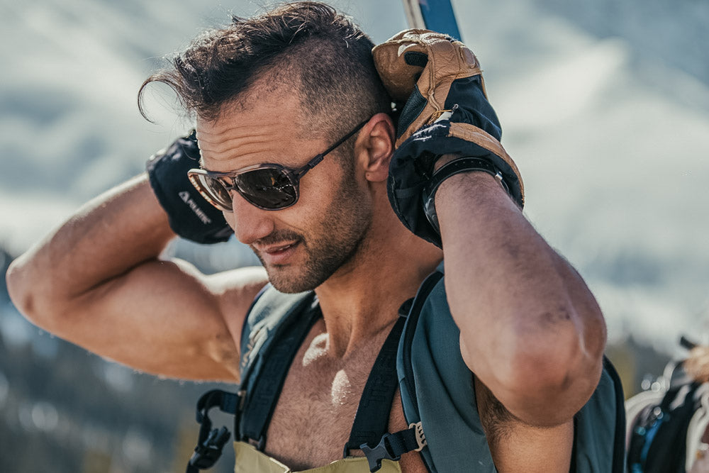 ember_grey Man hiking with a backpack and putting on his Ombraz leggero armless sunglasses with strap