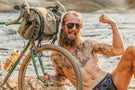 mattebrown_brown Tattooed man flex next to his bike wearing Ombraz classic armless sunglasses with strap
