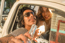 dolomite_charcoal_brown Couple in the front seat of a car sharing a drink wearing Ombraz armless sunglasses with cord
