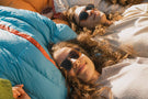 viale_dusk_brown Two people laying down smiling wearing Ombraz viale dusk brown armless strap sunglasses